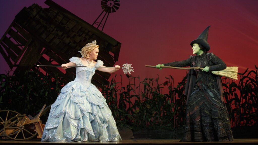 A promotional poster for the musical Wicked, featuring a green-skinned witch and her blonde counterpart against a dramatic background.